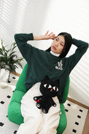 Model relaxing in green sweatshirt with black cat plush toy.