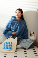 Model in blue hoodie with cat-and-fish patch, holding tote bag with cat.