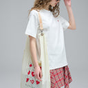 Model wearing a white t-shirt and red plaid skirt holding a cream-colored tote bag with heart designs.