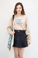 Model wearing a beige sweatshirt with text and a denim skirt, holding a blue bag against a white background
