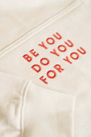 Close-up of red embroidered text "BE YOU DO YOU FOR YOU" on cream hoodie pocket