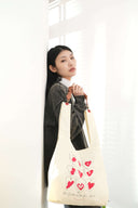 Model wearing Toby Black charcoal washed sweatshirt with hand-painted logo neckline, holding cream tote bag with red heart graphics
